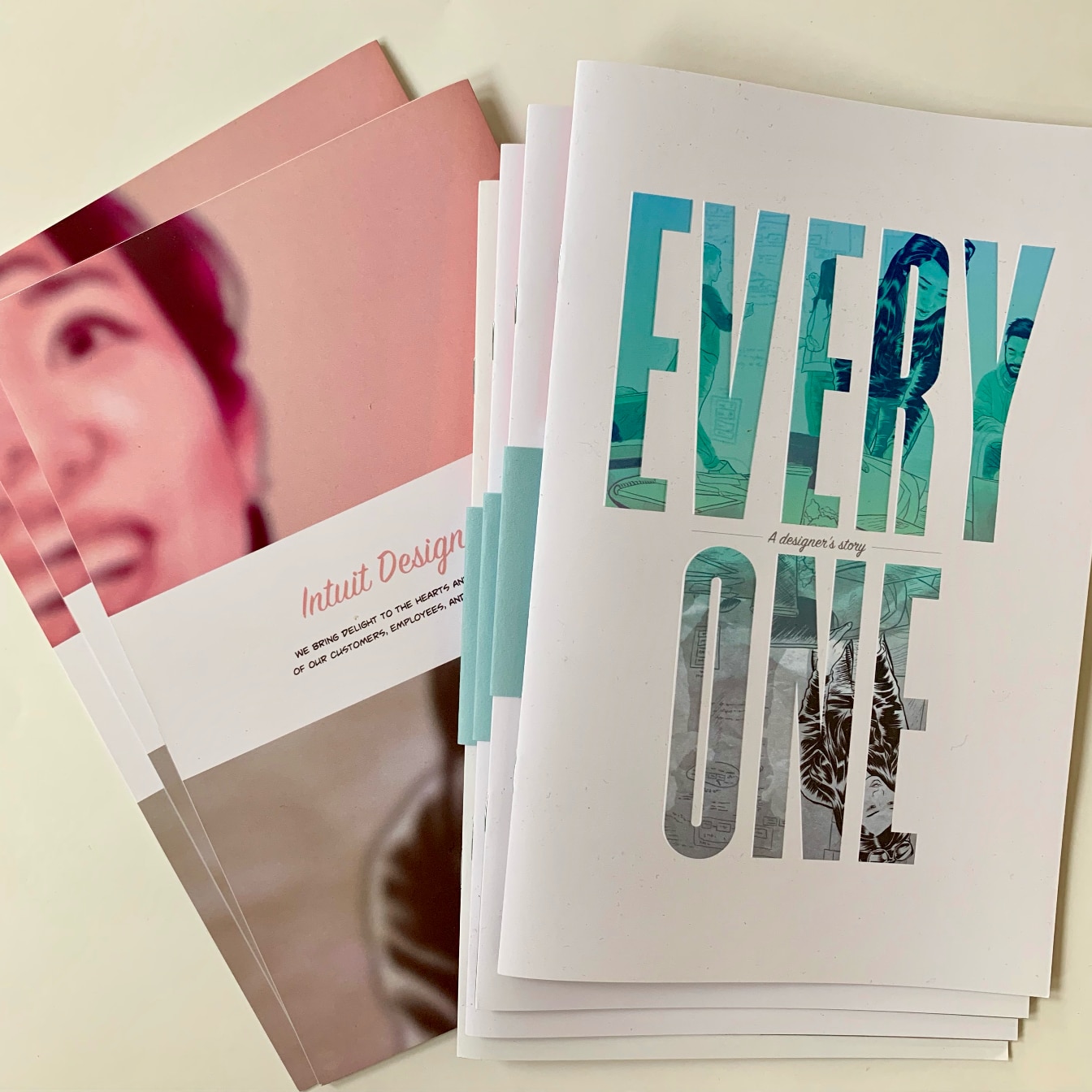 Printed copies of Everyone fanned out on a table. The front cover's large letters are filled with comic art, next to the pink back cover.